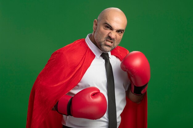 Бизнесмен-супергерой в красном плаще и боксерских перчатках смотрит вперед с сердитым лицом, готовым к бою, позирует, как боец, стоящий над зеленой стеной