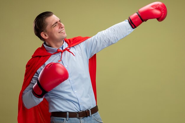 Super hero businessman in red cape and in boxing gloves making winning gesture with hand smiling confident ready to fight standing over light background