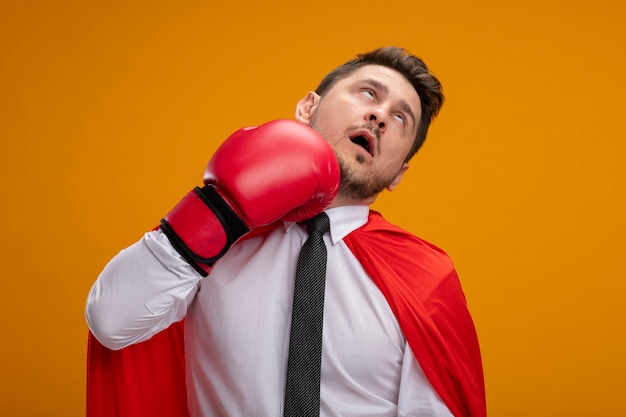 Super hero businessman in red cape and in boxing gloves punching himself standing over orange wall