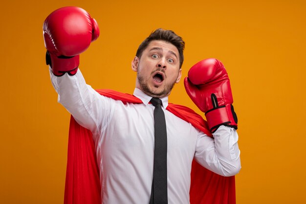 Super hero businessman in red cape and in boxing gloves  scared with hands out standing over orange wall