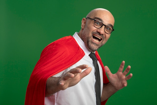 Super hero businessman in red cape and glasses happy and excited with arms out looking at front standing over green wall