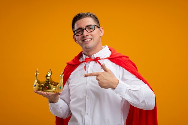 빨간 케이프와 안경에 검지 손가락으로 가리키는 왕관을 들고 슈퍼 영웅 사업가 오렌지 배경 위에 자신감이 서 웃고