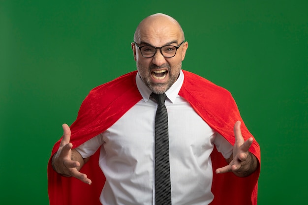 赤いマントと眼鏡をかけたスーパーヒーローのビジネスマンが、緑の壁の上に立って欲求不満と怒りを上げて叫び、叫んでいます