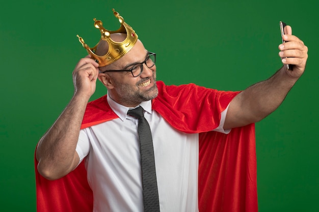 Super eroe uomo d'affari in mantello rosso e occhiali che indossa la corona facendo selfie utilizzando smartphone sorridente