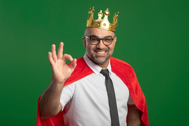 Foto gratuita super eroe uomo d'affari in mantello rosso e occhiali che indossa la corona guardando davanti sorridendo allegramente mostrando segno giusto in piedi verde muro bianco