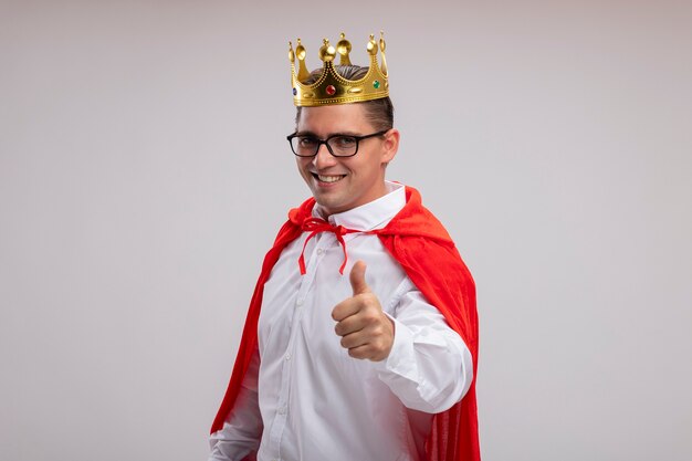 Super hero businessman in red cape and glasses wearing crown  smiling with happy face showing thumbs up standing over white wall