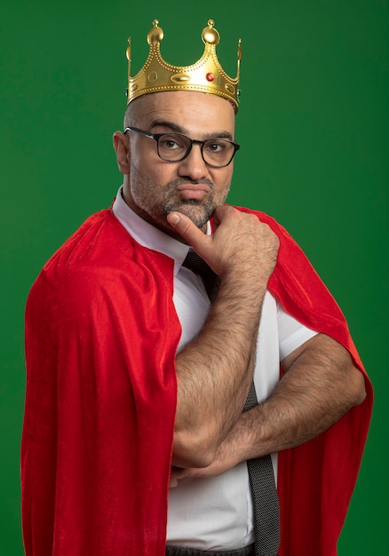 Super hero businessman in red cape and glasses wearing crown with hand on chin with confident serious expression 