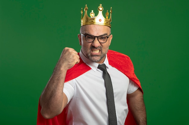 Super hero businessman in red cape and glasses wearing crown with serious frowning face clenching fist showing strength 