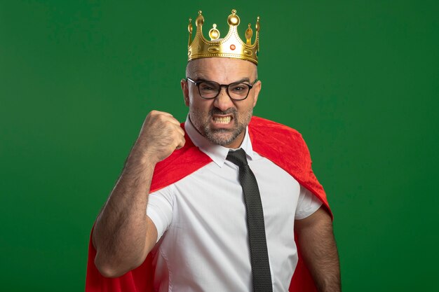 Super hero businessman in red cape and glasses wearing crown with serious frowning face clenching fist showing strength 