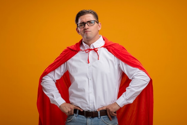 Super hero businessman in red cape and glasses  with serious face with arms at hip standing over orange wall