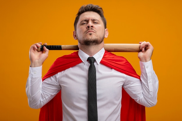 Foto gratuita uomo d'affari super eroe in mantello rosso che tiene la mazza da baseball dietro il collo guardando fiducioso in piedi sopra la parete arancione