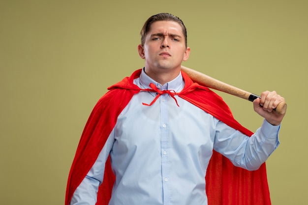 Super hero businessman in red cape holding baseball bat  with serious confident expression standing over light wall