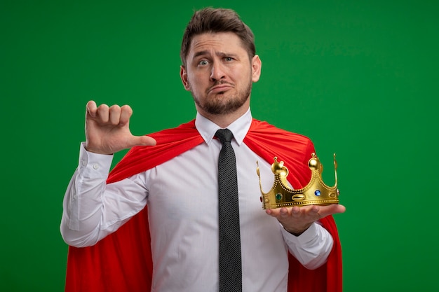 緑の背景の上に立って混乱していることを指している王冠を保持している赤いマントのスーパーヒーローの実業家
