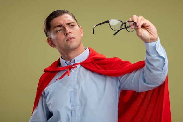 Super hero businessman in red cape holding glasses looking at them with interest standing over light background