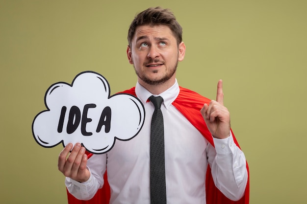 Super hero businessman in red cape holding speech bubble sign with word idea looking up showing index finger having new idea standing over green background