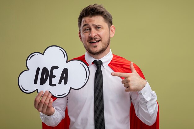 Super hero businessman in red cape holding speech bubble sign with word idea pointing with index finger at it smiling standing over green background