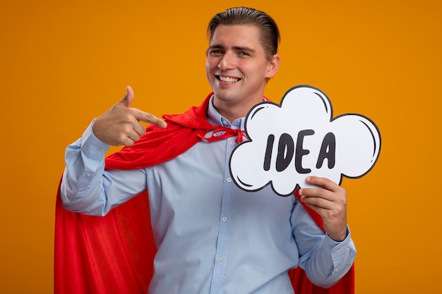 Super hero businessman in red cape holding speech bubble sign with word idea pointing with index finger at it smiling standing over orange background