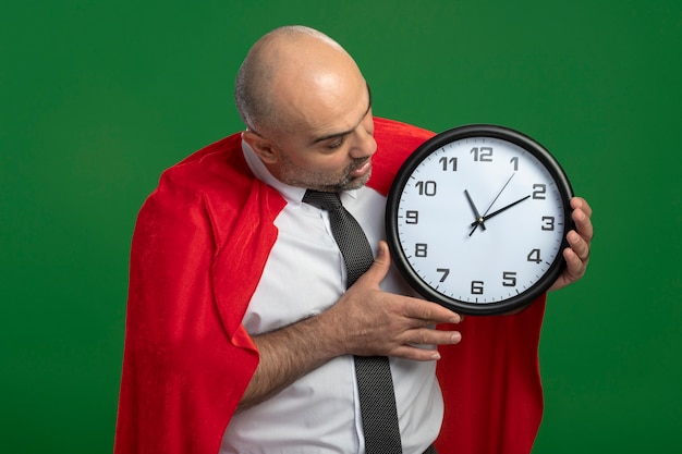 Бизнесмен супергероя в красном плаще держит настенные часы, глядя на него, будучи безумно пораженным и удивленным, стоя над зеленой стеной