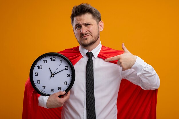 Super hero businessman in red cape holding wall clock pointing with index finger at it looking confused standing over orange wall