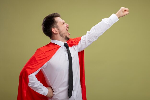 Super hero businessman in red cape keeping arm in flying gesture shouting ready to fight standing over green background