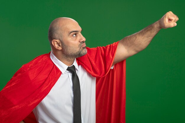 Бизнесмен супергероя в красном плаще смотрит в сторону, держа руку в летящем жесте, готовый помочь выглядеть уверенно, стоя над зеленой стеной
