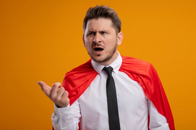 Super hero businessman in red cape looking aside with arm out as asking being confused standing over orange background