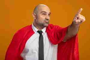 Foto gratuita uomo d'affari super eroe in mantello rosso che osserva da parte con espressione scettica che punta con il dito indice a qualcosa in piedi sopra la parete arancione