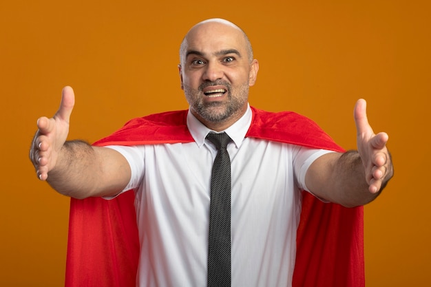 Super hero businessman in red cape looking at front smiling friendly making welcoming gesture with hands standing over orange wall