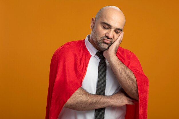 Super hero businessman in red cape looking at front with sad expression leaning head on his palm tired and bored standing over orange wall