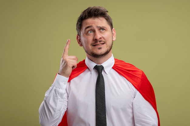 Super hero businessman in red cape looking up emotional showing index finger having great idea standing over green wall