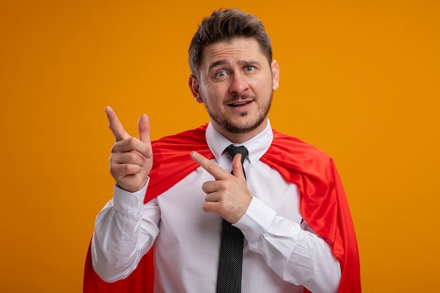 Super hero businessman in red cape pointing with index fingers to the side smiling standing over orange background