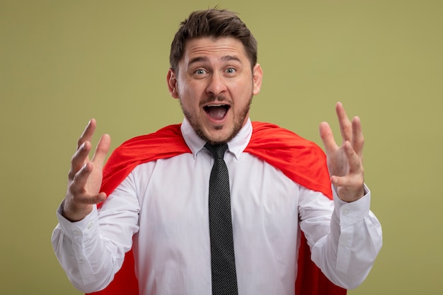 Free photo super hero businessman in red cape raising hands being amazed and surprised standing over green wall