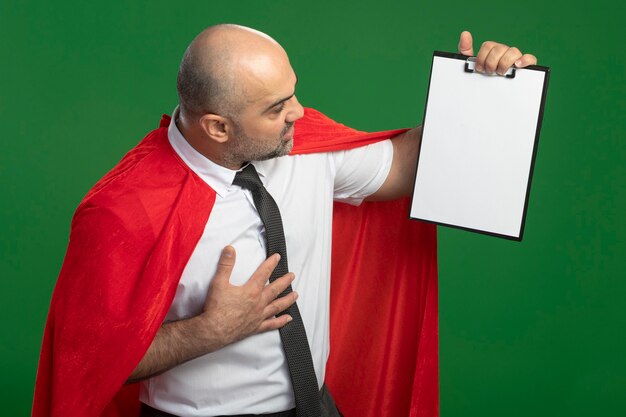 Free photo super hero businessman in red cape showing clipboard with blank pages looking at it intrigued