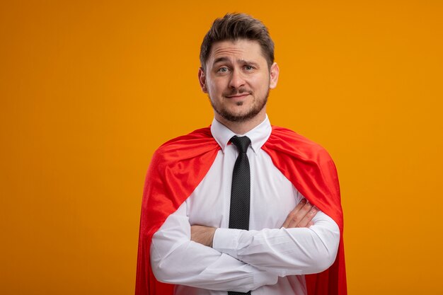 Super hero businessman in red cape  smiling confident with crossed arms on chest standing over orange wall