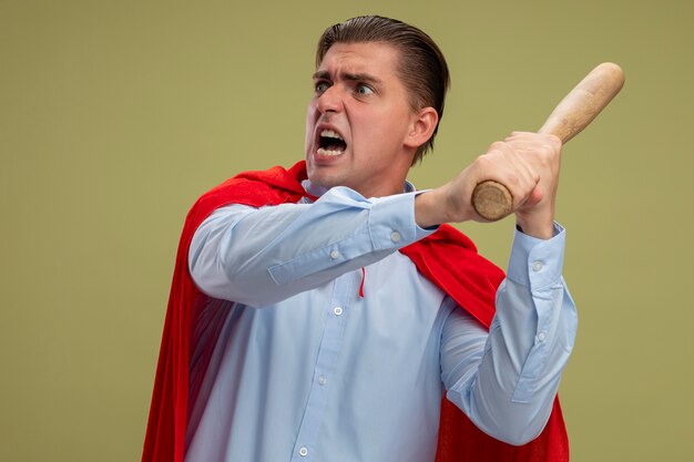 Super hero businessman in red cape swinging baseball bat with aggressive expression standing over light background