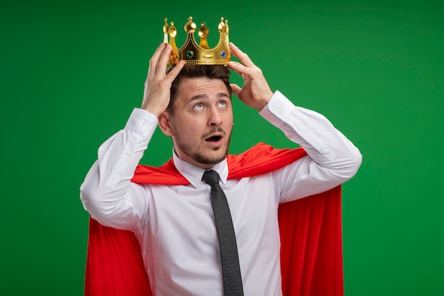 Super hero businessman in red cape touching his crown on his head being confused standing over green wall