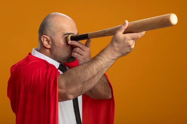 Free photo super hero businessman in red cape using baseball bat like spyglass standing over orange wall