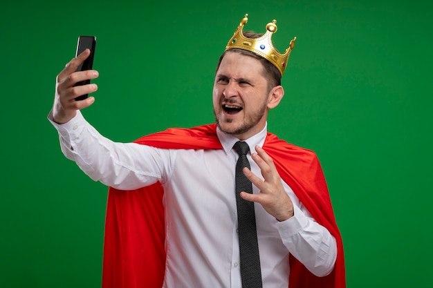 Супер герой бизнесмен в красном плаще в короне делает селфи с помощью смартфона, сходит с ума, злой, стоя на зеленом фоне