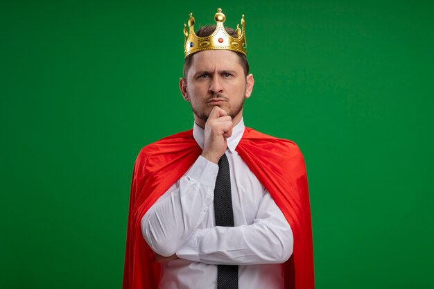 Super hero businessman in red cape wearing crown looking at camera with hand on chin with confident serious expression thinking standing over green background