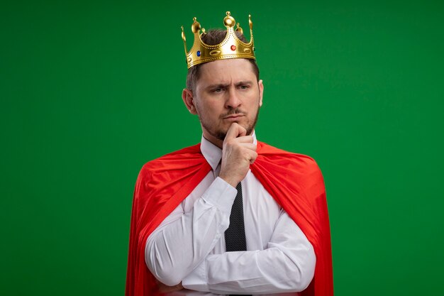 Супергерой бизнесмен в красном плаще в короне с рукой на подбородке с уверенным серьезным выражением лица, думающий, стоящий над зеленой стеной
