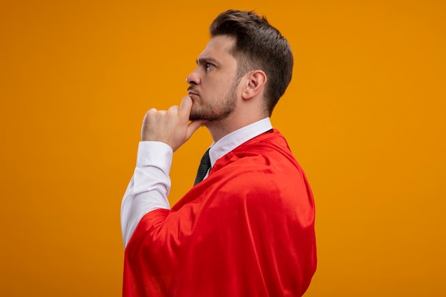 Super hero businessman in red capestanding sideways with pensive expression on face with hand on chin thinkingover orange wall