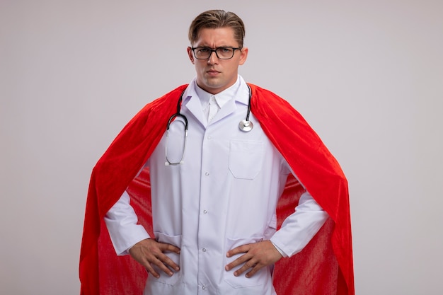 Super hero doctor man wearing white coat in red cape and glasses with stethoscope around neck looking at camera with serious confident expression with arms at hip standing over white background