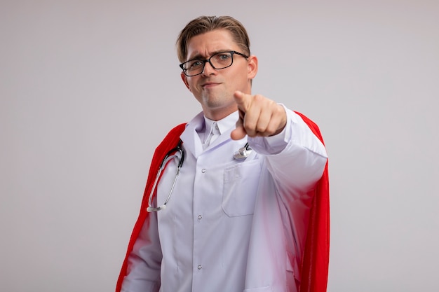 Super hero doctor man wearing white coat in red cape and glasses with stethoscope around neck pointing with index finger  smiling standing over white wall