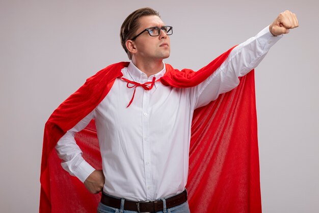 Человек-супергерой в красной накидке и очках смотрит в сторону с серьезным лицом, делая победный жест рукой, готовой помочь, стоя над белой стеной