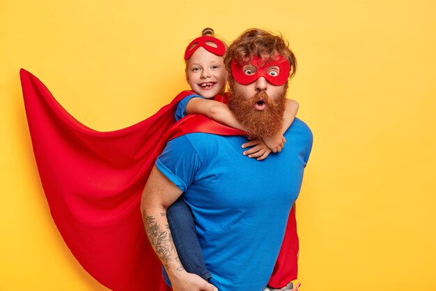 superheroes team ready to save our world. little kid girl rides back of her father superhero, pretend flying, wears red cape