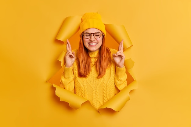 Superstitious redhead woman clenches teeth crosses fingers hopes dreams come true wears yellow hat and sweater breaks through paper hole. Ginger millennial girl believes in good luck. Wish concept