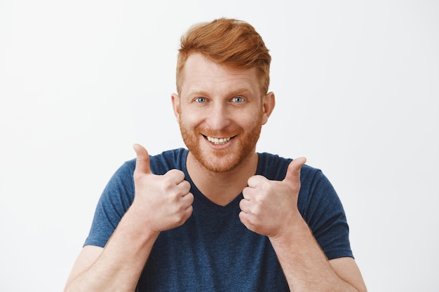Supportive and attractive adult redhead male with bristle liking great plan raising hands, showing thumbs up, smiling joyfully