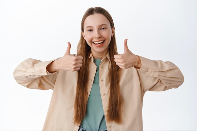 Supportive girl shows thumbs up, smiling and say yes, nod in approval, encourage you, praise for good choice, nice work, well done gesture, standing over white background.