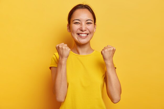 Supportive good looking triumphs with success, raises clenched fists, smiles happily, has eastern appearance, happy finally gaining goal, glad to fullfil dream, dressed casually poses over yellow wall