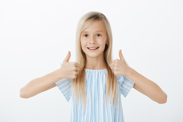Supportive little friend wants to cheer mates. Happy positive cute girl with blond hair, raising thumbs up and giving approval, smiling broadly while liking great idea, standing over gray wall